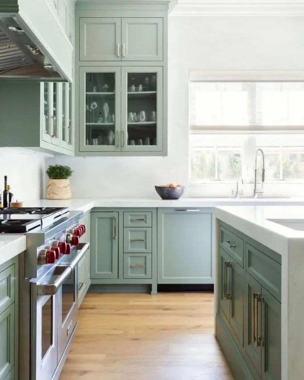 a beautiful sage green kitchen with shaker and glass front cabinets, white stone countertops and a backsplash feels airy