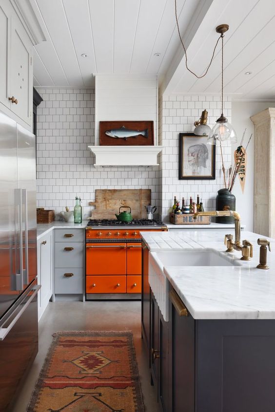 a bright eclectic kitchen with an orange cooker, navy and grey cabinets, white stone countertops and catchy artworks