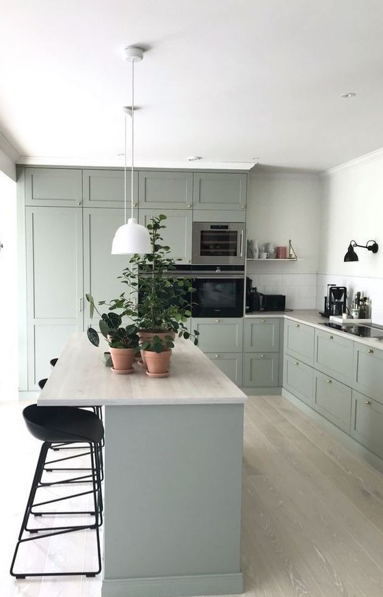 a lovely and soothing sage green kitchen with shaker cabinets, a kitchen island, black stools, pendant lamps and a white tile backsplash