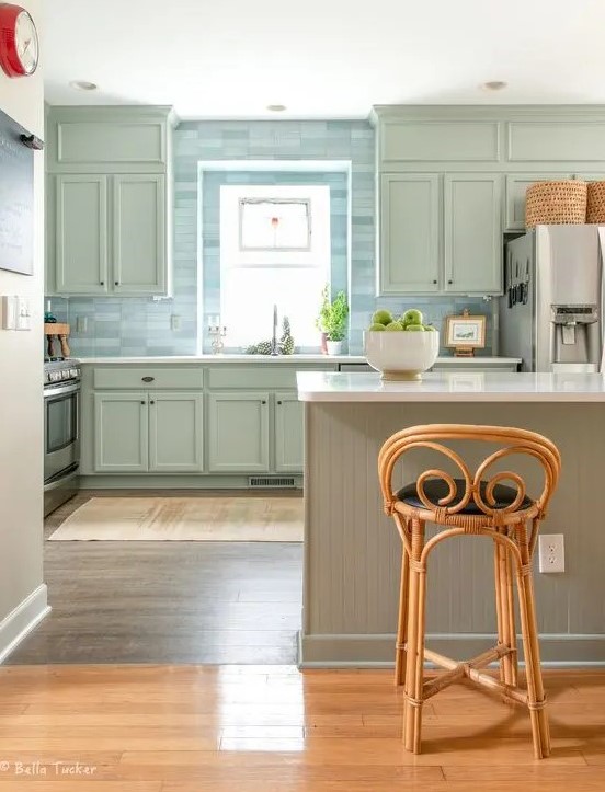 a lovely sage green kitchen with shaker cabinets, a blue skinny tile backsplash a slatted kitchen island and vintage rattan stools