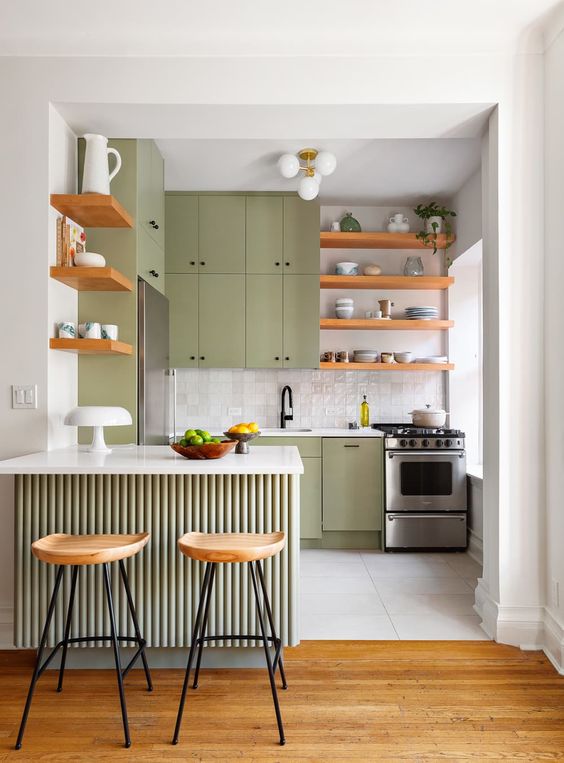 a pretty green kitchen with a white tile backsplash, stained shelves, a fluted kitchen island and tall stools