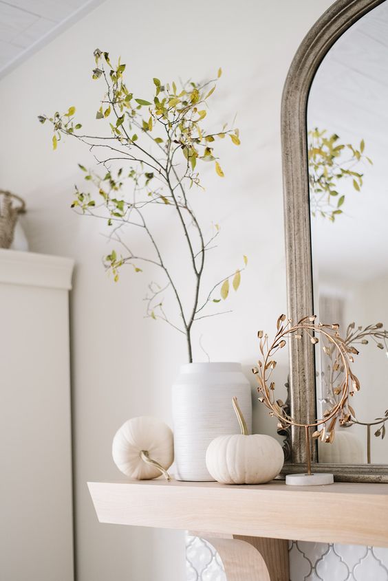 Simple Scandinavian mantel decoration with white pumpkins, a white vase with green branches and a gold wreath on a stand