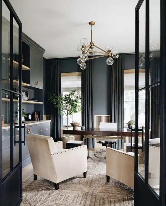 a beautiful soot-colored home office with cabinets, a stained desk, cream chairs, a chic chandelier and lots of greenery