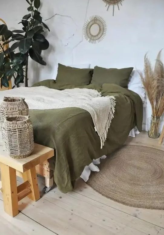 A boho bedroom in neutral tones features a bed with olive green linens, a wooden bench and woven candle holders, pampas grass, potted plants and broken mirrors