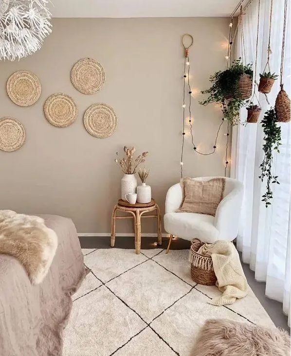 a boho bedroom with taupe walls, a bed, a cream chair, a rattan stool, hanging flower pots, decorative woven plates and layered rugs