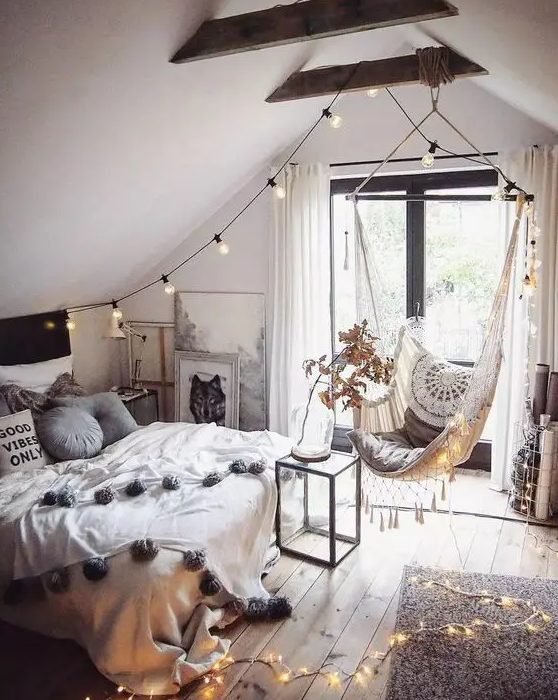 A boho teen bedroom in the attic features a bed with neutral linens, a woven hanging chair, lights, wooden beams and artwork