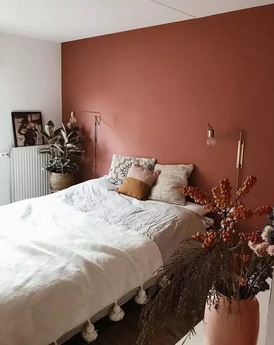 a striking bedroom with a terracotta accent wall, bed with neutral linens, gold sconces and potted plants