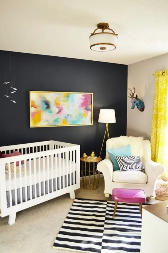 A bright, mid-century modern nursery with a black accent wall, white furniture, layered rugs, a bright piece of art and some pillows, and a pink ottoman
