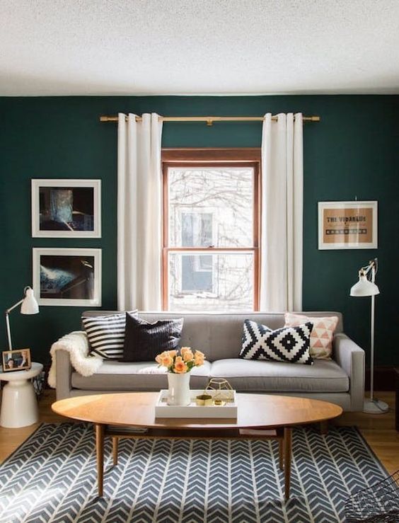 a pretty little living room with dark green walls, a gray sofa with printed cushions, a printed rug, an oval table and some artwork