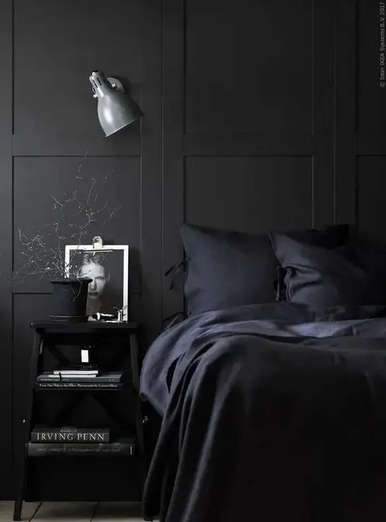 A chic, Nordic-inspired bedroom with black paneling, dark blue linens and black furniture, as well as a wall light