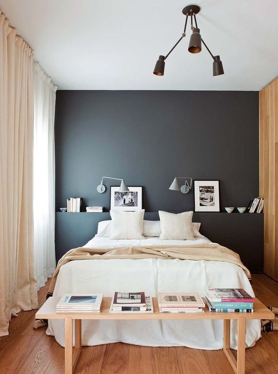 a chic, modern bedroom with a black accent wall, a bed with neutral linens, a bench with books and a chandelier