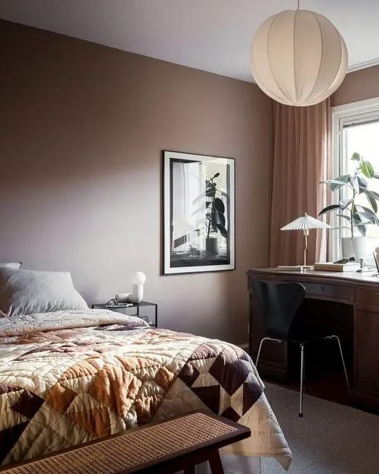 a cozy bedroom with a brown accent wall, a bed with printed linens, a dark stained desk, a black chair and some plants