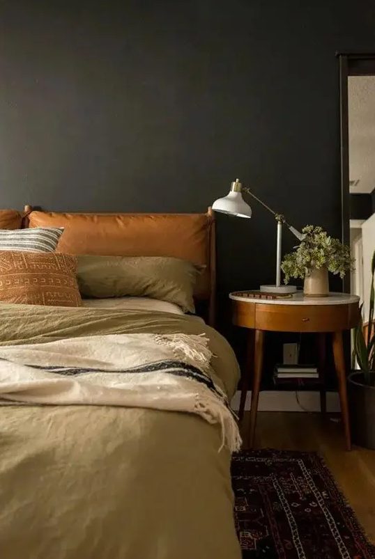 a cozy, atmospheric bedroom with sooty walls, an amber leather-covered bed, neutral linens, a chic bedside table and a printed rug