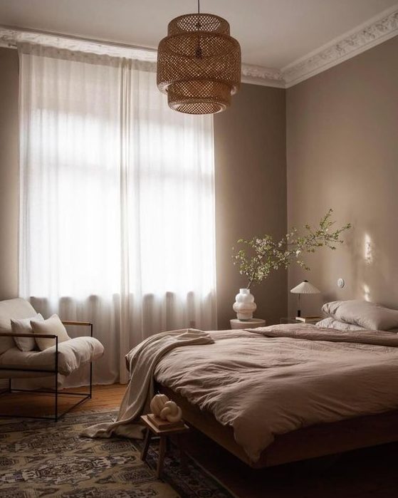 a dreamy light brown bedroom with a bed with neutral linens, a printed rug, a woven pendant lamp and a white chair