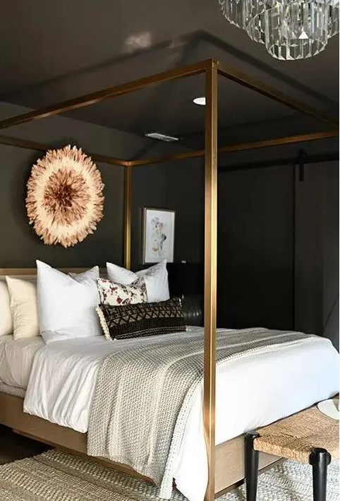a glamorous, moody bedroom with sooty walls and ceiling, a gold four-poster bed with neutral linens, a woven bench and a crystal chandelier