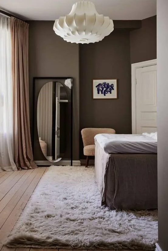 a laconic bedroom with brown walls, a high bed, a neutral carpet, a floor mirror, mauve curtains and a lamp