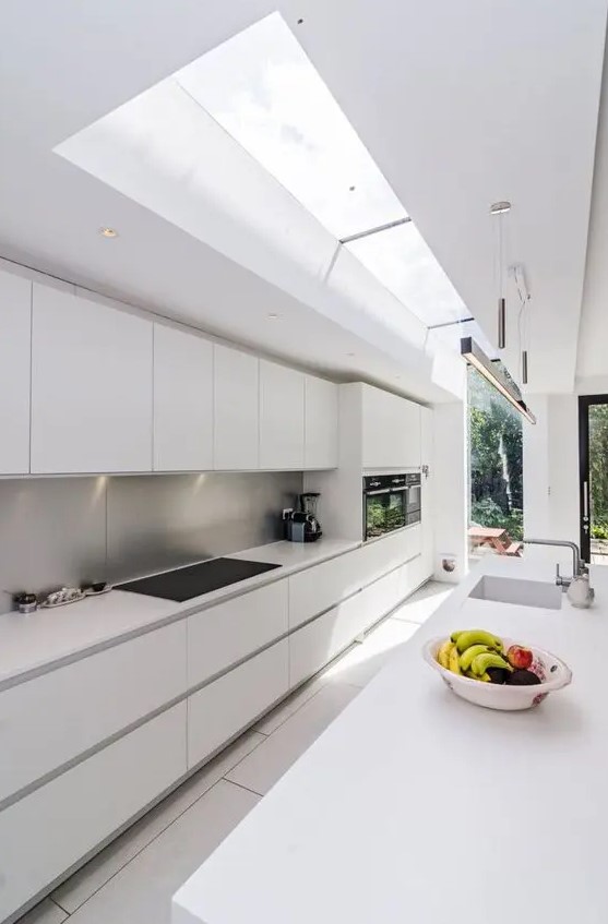 A long, narrow, minimalist white kitchen with a metal backsplash and skylights that let in lots of light