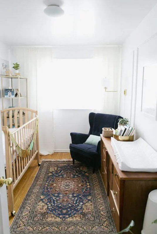 A small, mid-century modern nursery with a stained crib, stained dresser, navy wingback chair, printed rug and high shelf
