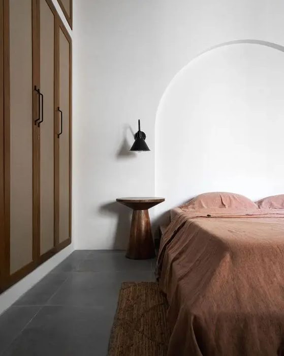 a minimalist, earthy bedroom with a built-in wardrobe, a bed with rust linens, a metal bedside table and a black wall lamp