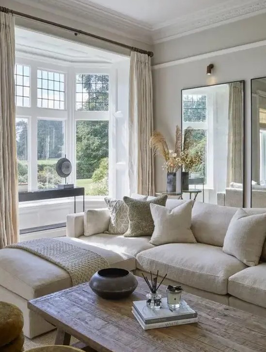A modern, country living room in neutral tones, with mirrors, a cream corner sofa, a bay window, a wooden table and cool textiles