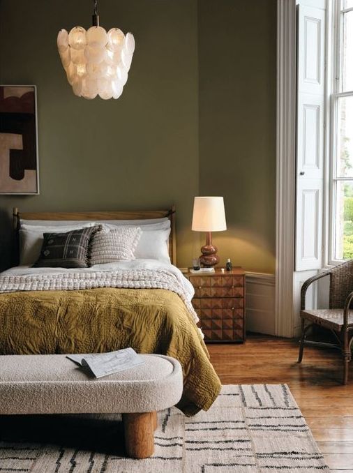 A modern earth toned bedroom with green walls, a stained bed with statement linens, a bouclé bench and a geometric bedside table