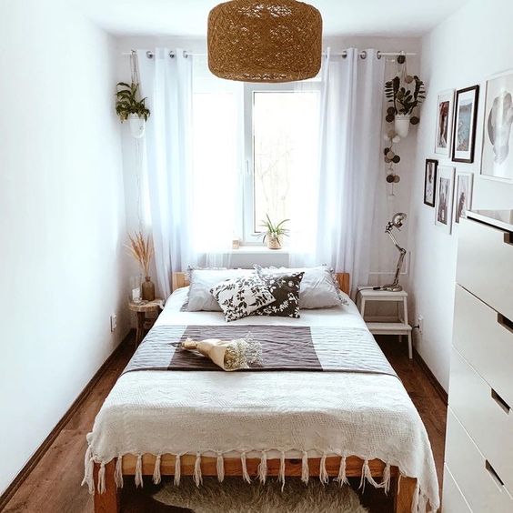 a modern, narrow bedroom with a bed with printed linens, mismatched bedside tables, potted plants, a gallery wall and a chest of drawers
