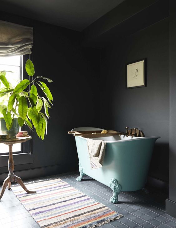 a moody, sooty bathroom with an aqua blue freestanding bathtub, a colorful rug, a potted plant, books and some art