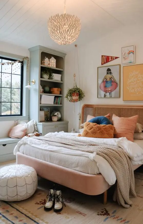 A boho bedroom in muted colors with a pink bed with a rattan headboard, a light green storage area with a windowsill and a chandelier