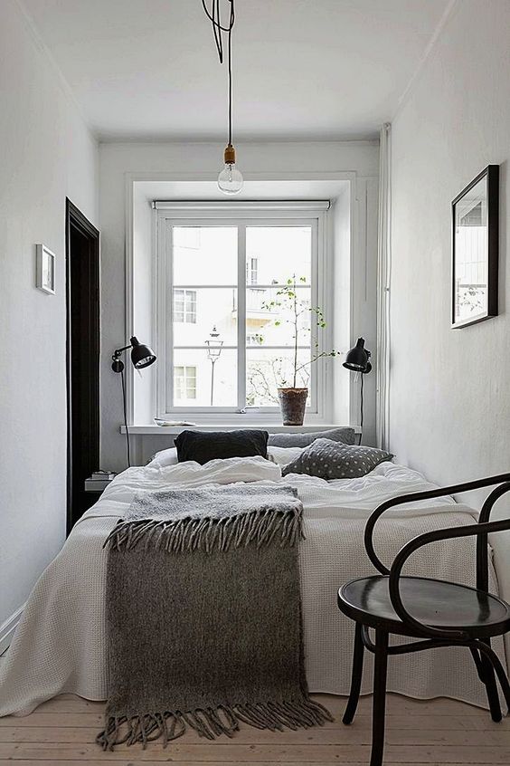 a narrow Scandinavian bedroom with a bed, black sconces and a black chair, some greenery and a piece of art