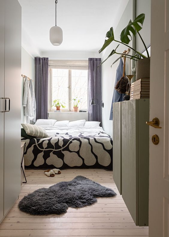 a narrow bedroom with a bed and printed linens, mauve curtains, a rug, a green cupboard, and potted plants and books
