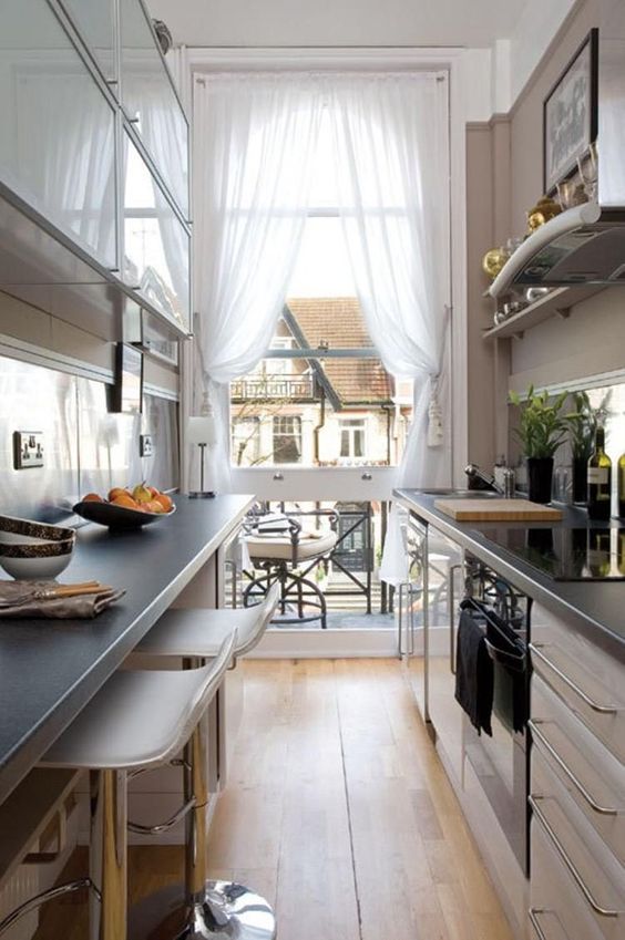 a narrow kitchen with neutral cabinets, black countertops, high stools, ledges and shelves and a view through the balcony entrance
