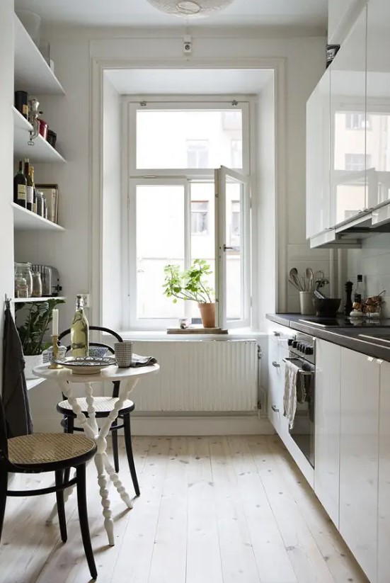 a narrow kitchen with open shelving, sleek white cabinets, black countertops, a small table and wicker chairs, and potted plants