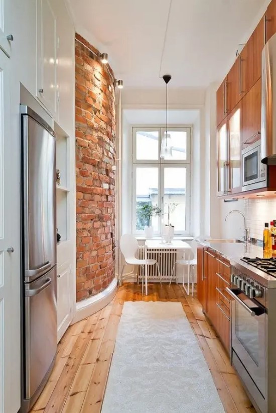 a narrow kitchen with richly stained cabinets, built-in appliances, a brick wall, a white dining set and a stained floor