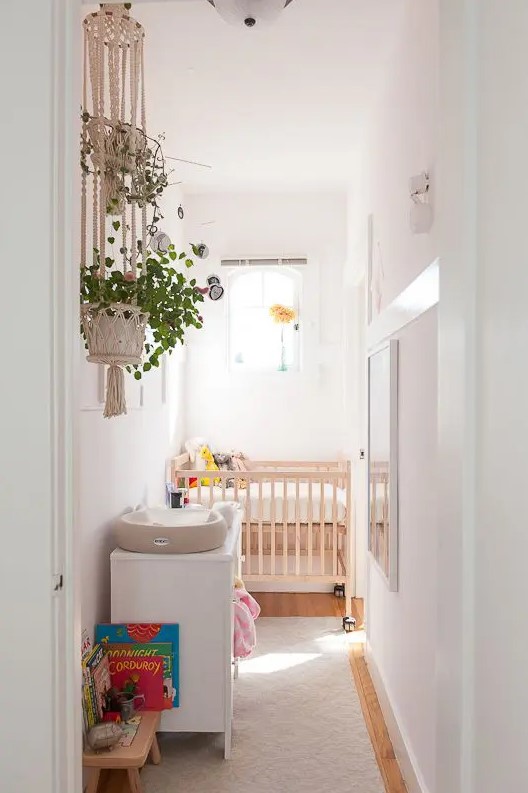 a narrow white children's room with a stained cot, a white dresser, some windows and artwork, green plants and toys