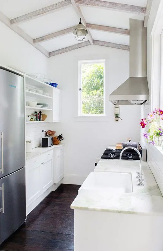 a narrow white kitchen with shaker-style cabinets, white stone countertops, windows, bright flowers and a refrigerator