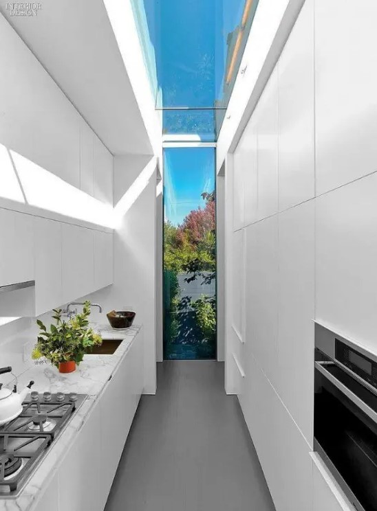 a narrow white kitchen with sleek cabinets, built-in appliances and a skylight leading into a window onto the garden for natural light and views