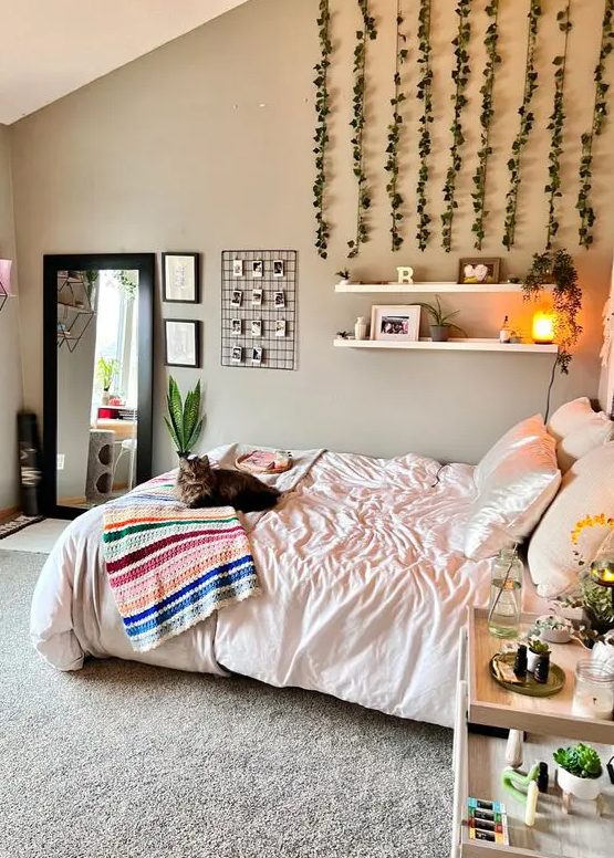 a neutral boho bedroom with gray walls, a bed with neutral linens, open shelves, hanging greenery and a bedside table with flowers