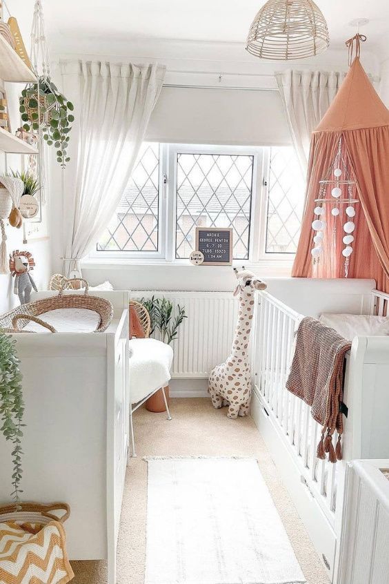 A neutral boho nursery with a large white crib, changing table, chair, bookshelves, coral canopy and pretty decor