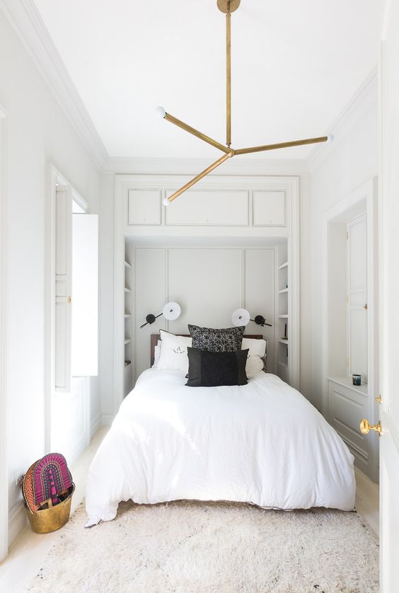 a neutral, narrow bedroom with built-in shelves on the headboard, a bed with black and white linens, a chandelier and some bright decor