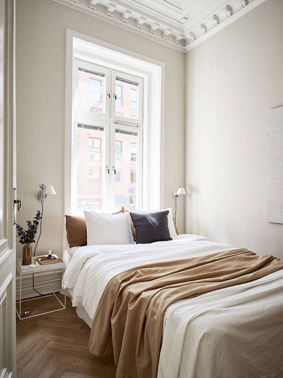 a neutral, narrow bedroom with moldings, a bed with muted-colored linens, a few nightstands and sconces