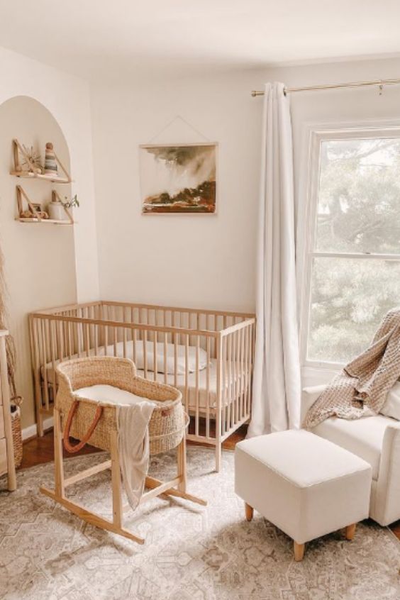 a neutral nursery with a stained crib, bassinet, white chair and ottoman, printed rug, bookshelves and artwork