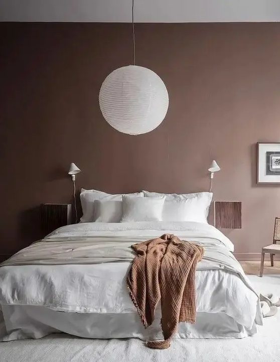 a pretty bedroom with brown walls, a bed with neutral linens, floating nightstands, a pendant lamp and a chair
