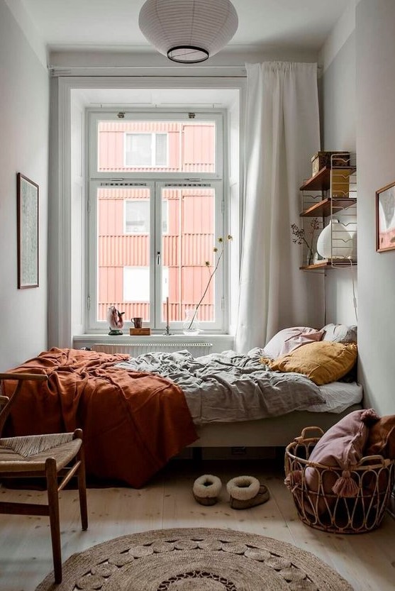 a pretty boho bedroom with a shelf, a bed with mustard and rust linens, a basket with pillows, a jute rug and a chair
