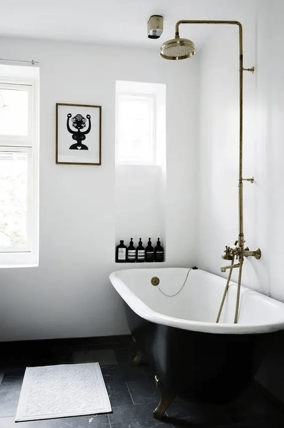 A calm white bathroom with niches for storage, a black clawfoot tub, brass fixtures, and fun artwork is cool