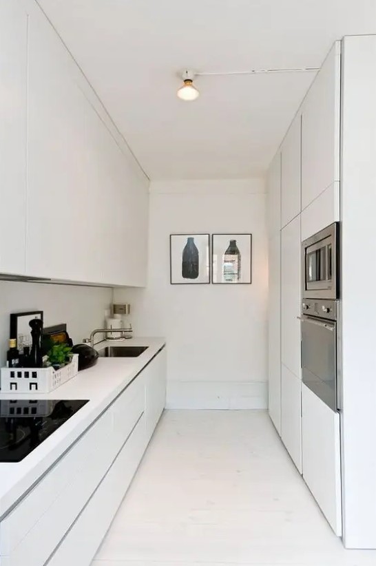 A sleek, narrow white kitchen with minimalist cabinets, built-in appliances and some artwork is amazing