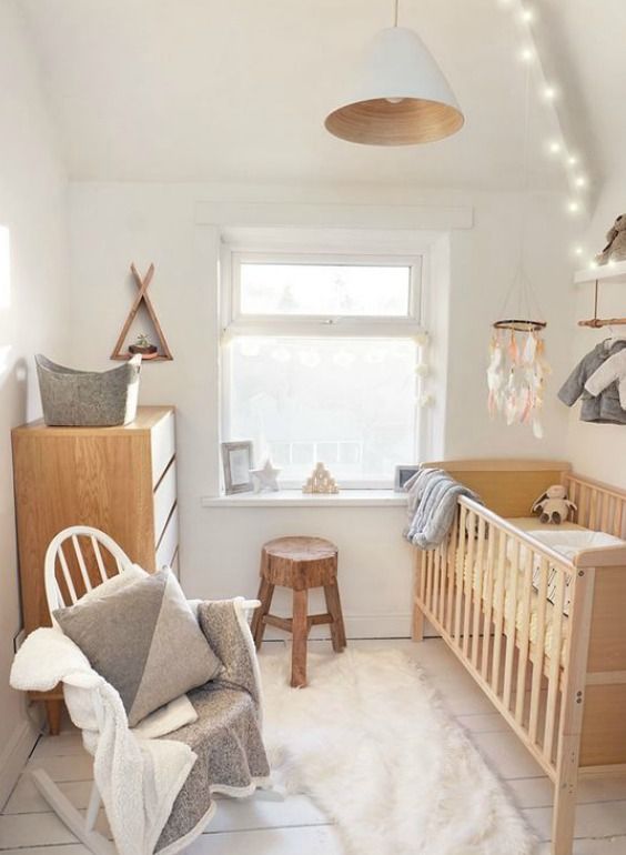A small Scandinavian nursery with a light stained crib, white rocking chair with pillows, dresser, stool and pretty decorations