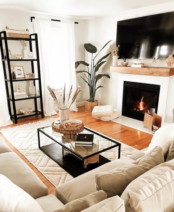 a small and inviting living room with a fireplace, neutral seating area, shelf, coffee table, potted plants and cushions