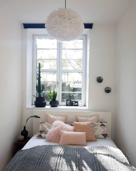 a small and narrow bedroom with a white bed, pink and printed bed linen, a single bedside table and some potted plants