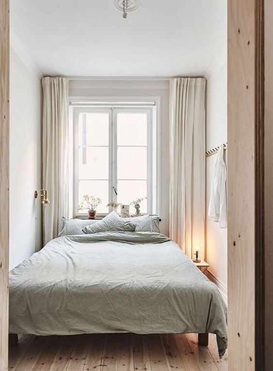 a small and narrow neutral bedroom with a bed with light green linens, a bedside table with a lamp and neutral curtains for peaceful sleep