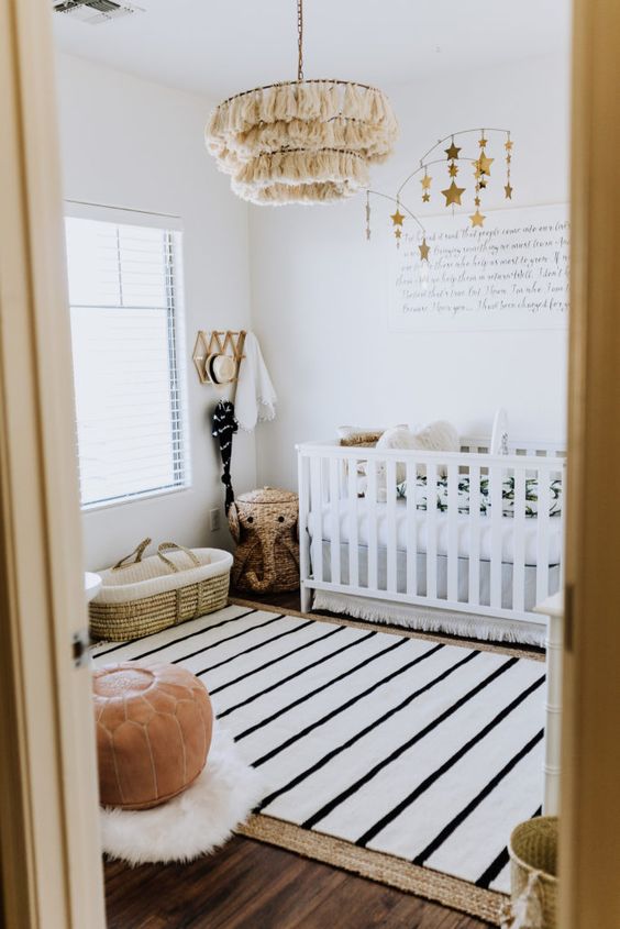 a small boho nursery with layered rugs, a white crib with printed linens, a leather stool, a tassel chandelier and pretty decorations